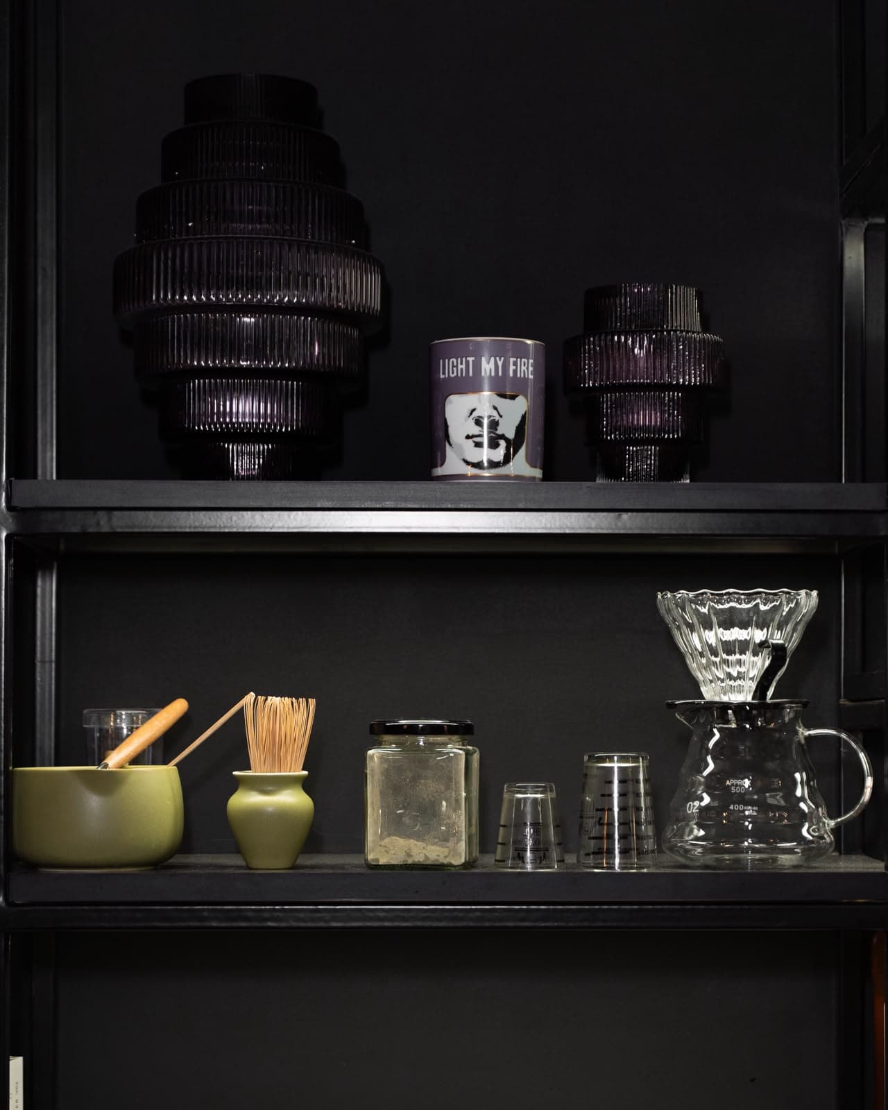 Black shelving in a Sella store: ribbed plum glass vessels and a candle above, a matcha bowl, jars and pour-over glassware below