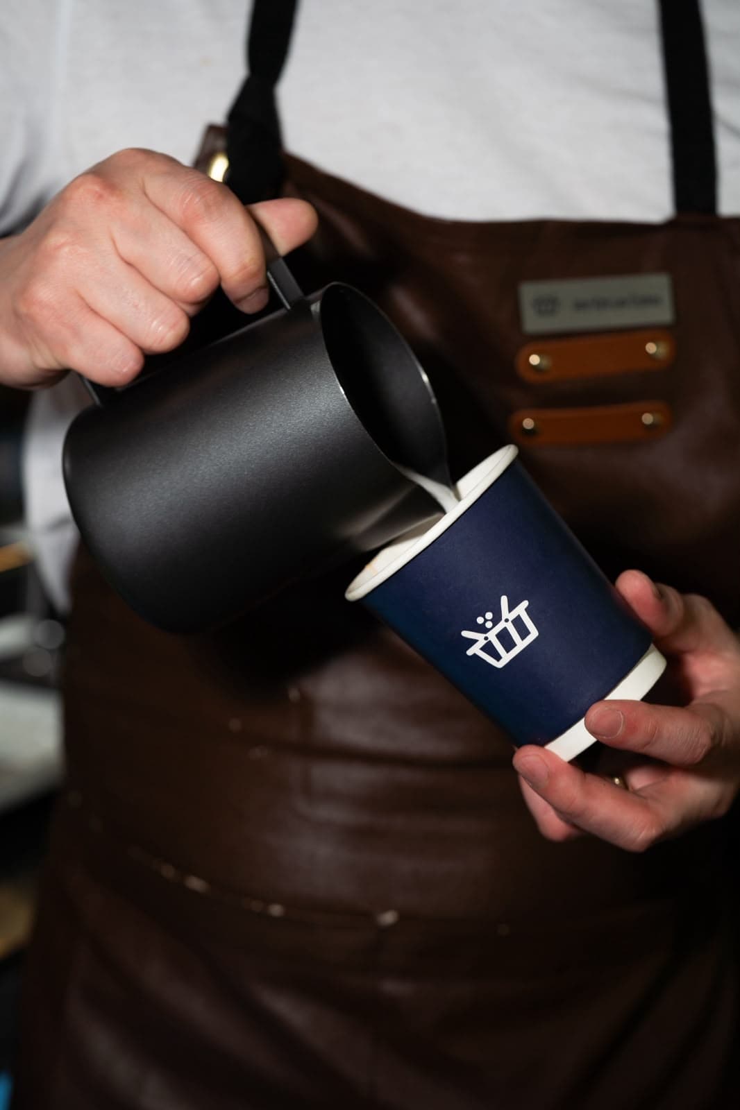 A barista in a leather apron pouring steamed milk from a black pitcher into a navy Sella-branded cup