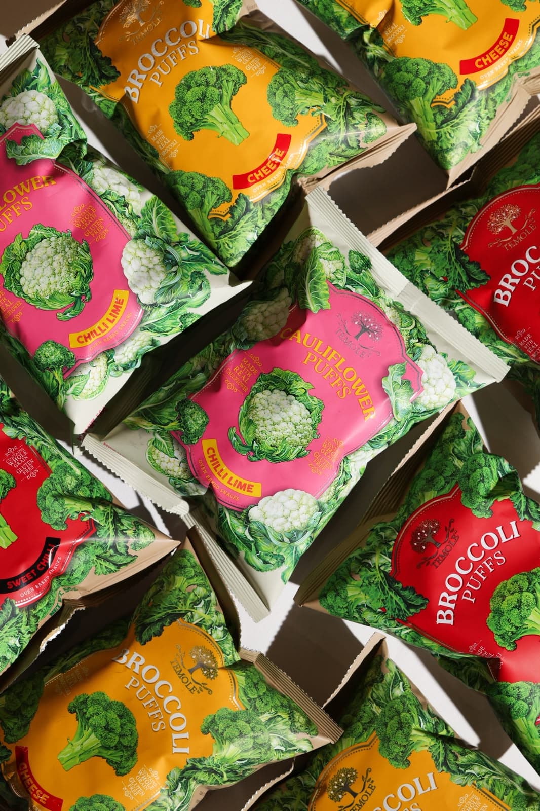 Packets of broccoli and cauliflower puffs in yellow, pink and red, scattered on a white surface